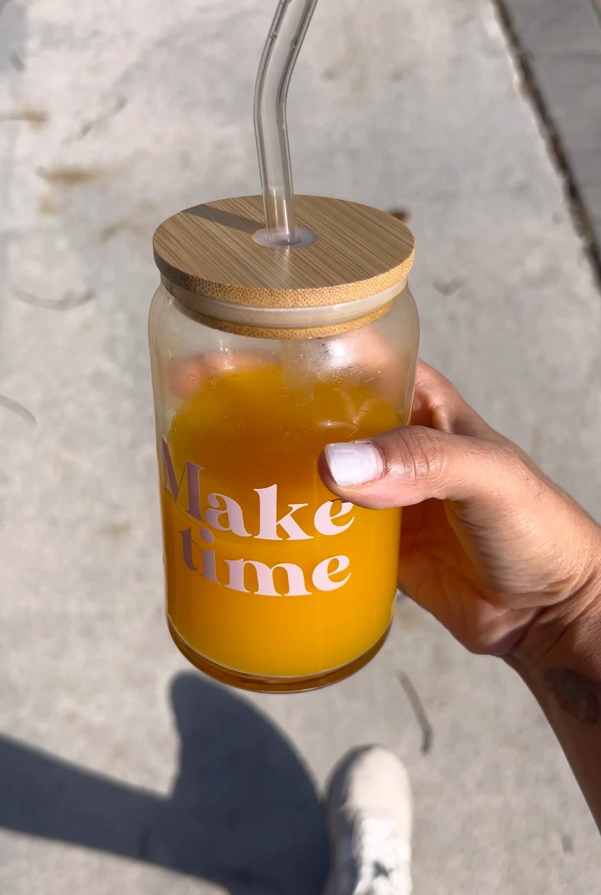 Hand holding a glass jar with a wooden lid and straw, filled with orange juice and text 'Make time'.