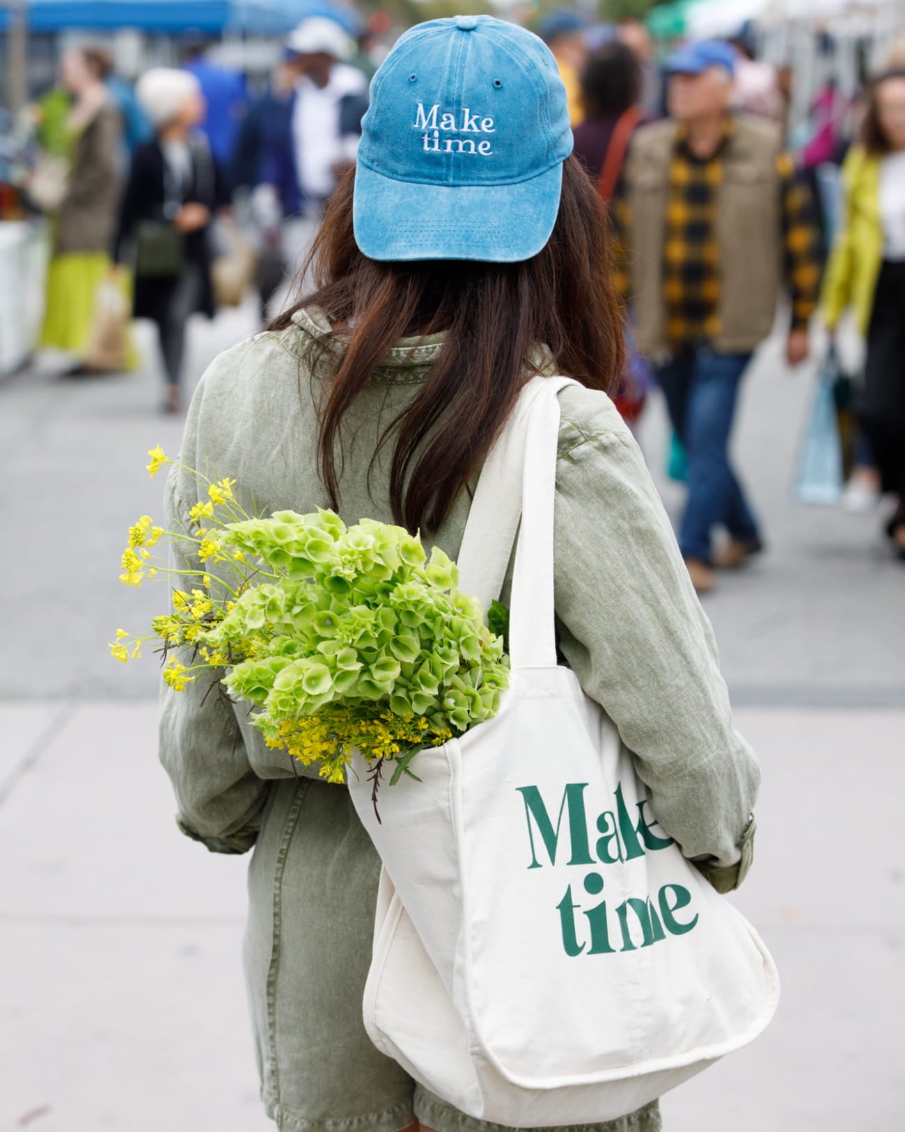 Person wearing a blue cap and holding flowers, with a tote bag featuring 'Make time' text, in an urban setting.